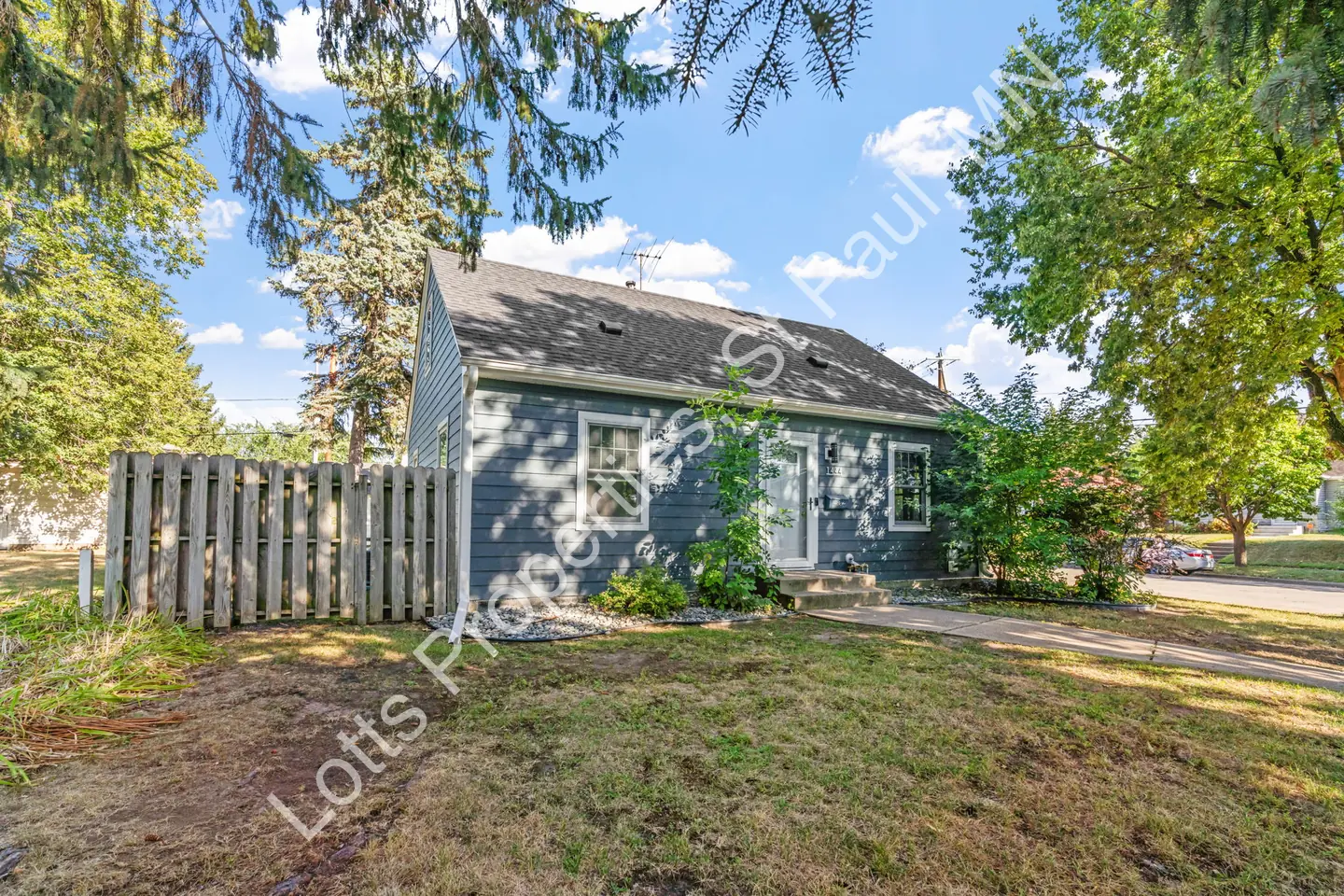 Side-angle view of the blue home's front with a wood privacy fence and mature trees.