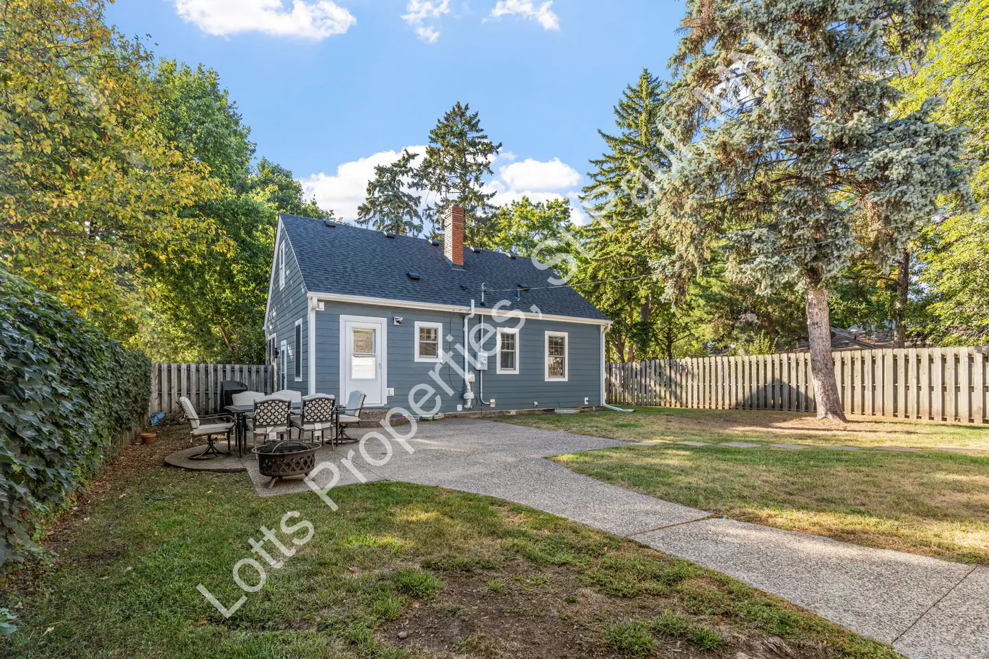 Back exterior showing a patio with table and fire pit beside the blue home and a fenced yard.