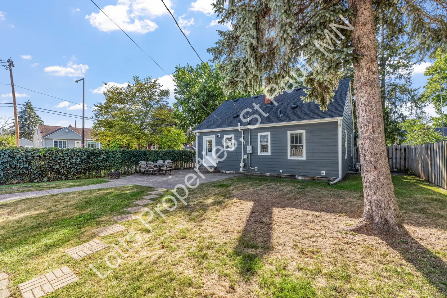 Fenced backyard with a mature spruce tree, paver path, and patio behind the blue home.