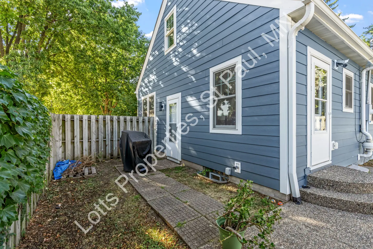 Side entry of the blue home with a white door, steps, and a covered grill along a wood fence.